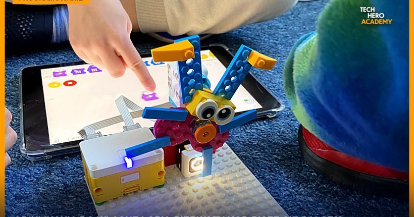 Dziecko wskazuje palcem kod blokowy na ekranie tabletu, obok robot zbudowany z klocków LEGO na zajęciach Tech Hero Academy