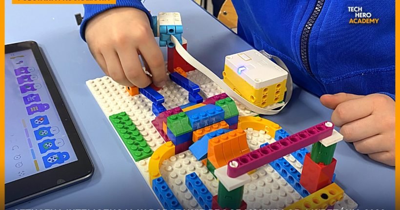 Uczeń buduje przy stole model z klocków LEGO z modułem elektronicznym, obok tablet z aplikacją do programowania blokowego